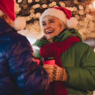 A cheerful grandmother and friend in Santa hats talk and laugh outdoors under twinkling Christmas lights, holding hot coffee cups during a festive holiday moment.