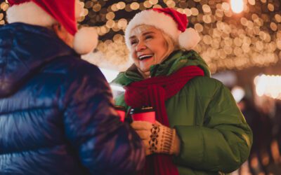 A cheerful grandmother and friend in Santa hats talk and laugh outdoors under twinkling Christmas lights, holding hot coffee cups during a festive holiday moment.